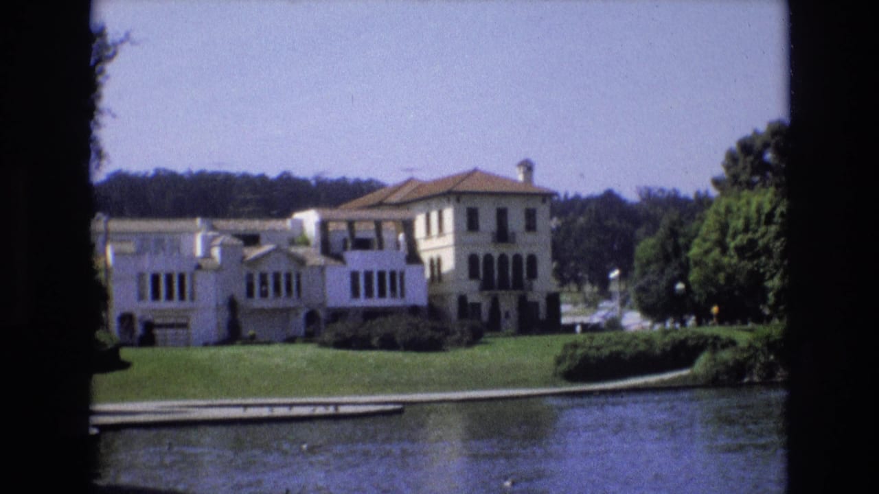 Large white house and lawn beside pond water under a bright sky.