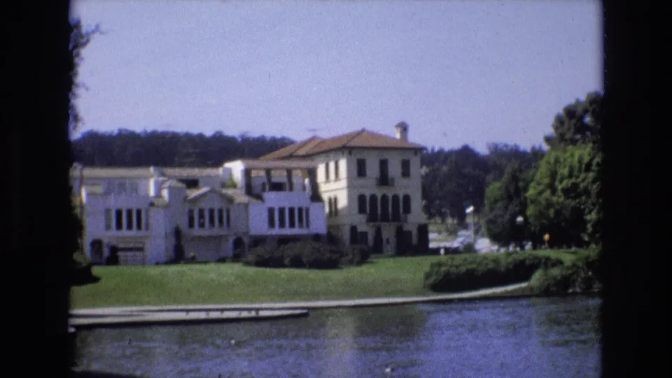 1973 San Francisco 8mm frame with the white stucco-style mansion centered beyond pond water, framed by lawn, shrubs, and trees.