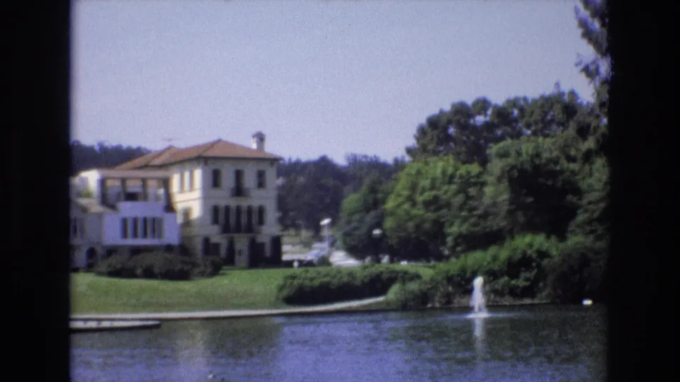 1973 San Francisco 8mm frame showing the white mansion partly obscured by trees, with pond water and a small fountain in the foreground.