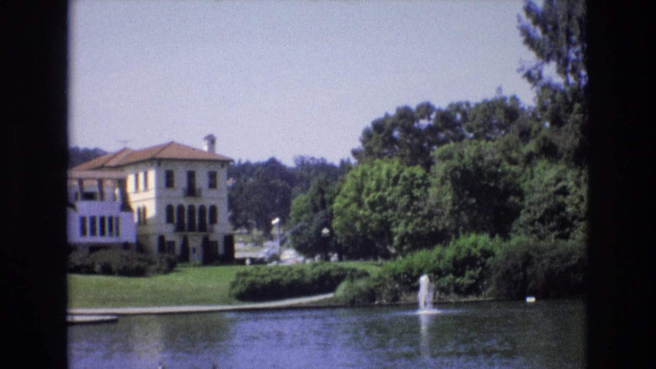 1973 San Francisco 8mm frame showing a pale Mediterranean-style lakeside building, red-tiled roof, green lawn, trees, and a small fountain spraying in the pond.