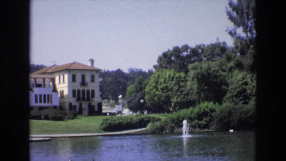 1973 San Francisco 8mm frame showing a pale Mediterranean-style lakeside building, red-tiled roof, green lawn, trees, and a small fountain spraying in the pond.