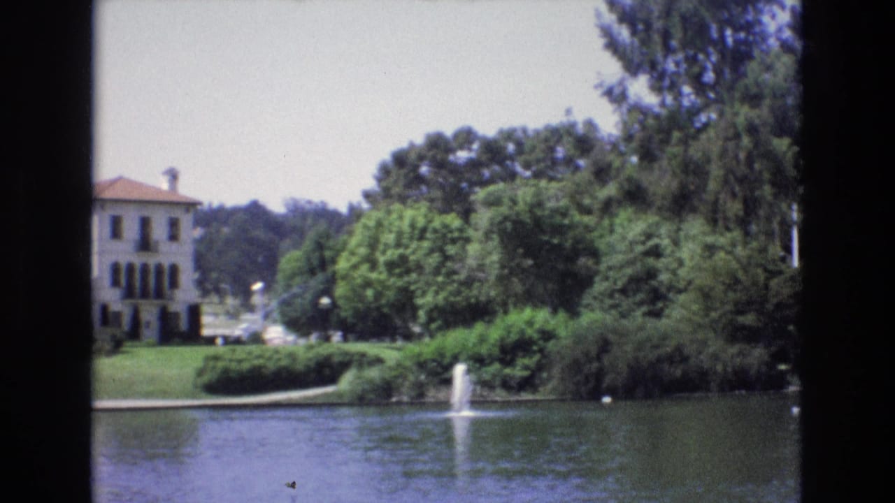 1973 San Francisco 8mm frame with the lakeside building partly obscured by trees, fountain spray in the pond, trimmed greenery, and bright sky.