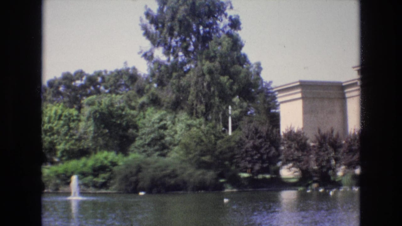 1973 San Francisco home-movie frame showing a pond with a small fountain, dense greenery, and a partial view of a pale classical landmark building beyond the water.