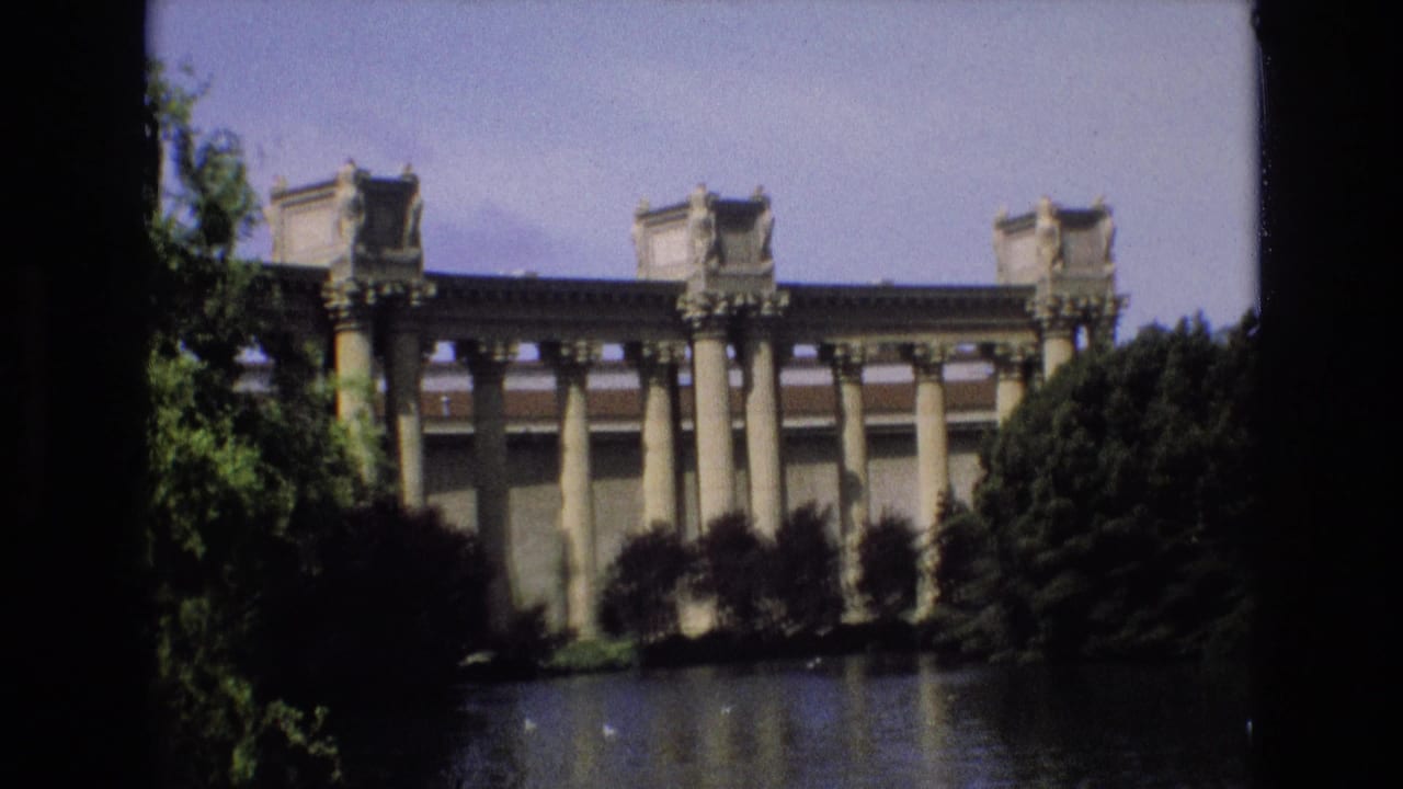1973 San Francisco home-movie frame showing a grand classical colonnade across water, with trees and shrubs partly obscuring the landmark facade.