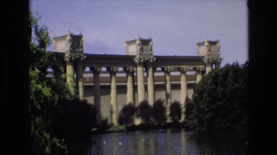 1973 San Francisco home-movie frame showing a grand classical colonnade across water, with trees and shrubs partly obscuring the landmark facade.
