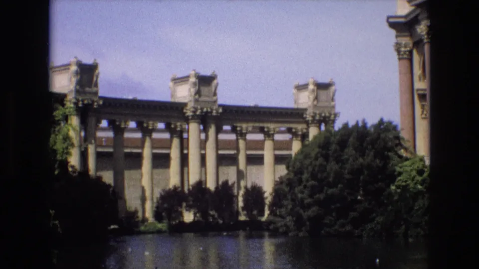 1973 San Francisco home-movie frame with a wide view of a classical colonnaded structure, pond reflections, surrounding trees, and a partial foreground architectural edge.