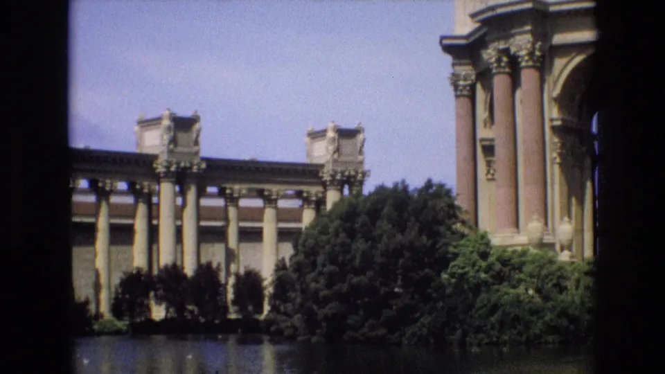 1973 San Francisco home-movie frame showing a classical colonnaded landmark across the pond, bordered by trees and a cropped architectural element at the right.