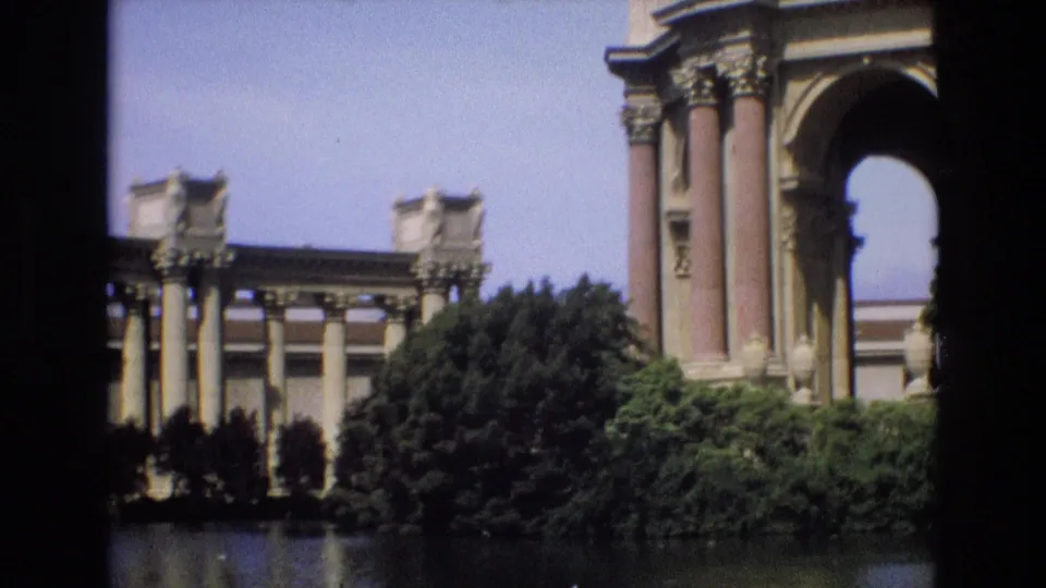 1973 8mm view of the Palace of Fine Arts in San Francisco, with columns, an arch, trees, shrubs, and water framed by dark film borders.