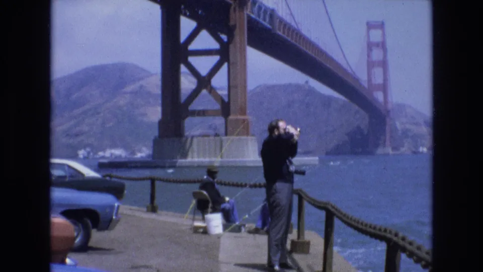 1973 San Francisco home-movie frame of a visitor using binoculars near parked cars and a railing, with the Golden Gate Bridge across the water.