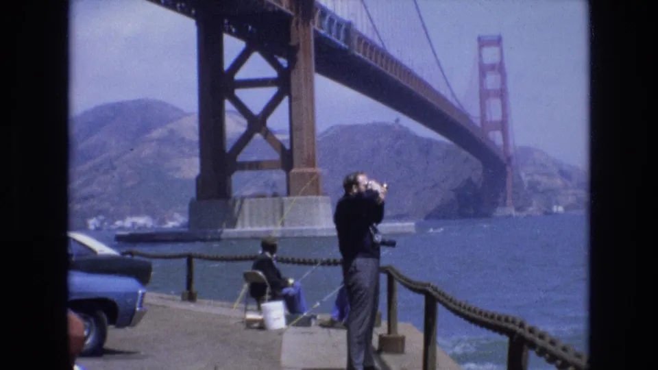 1973 San Francisco 8mm frame showing a visitor with binoculars at a waterfront railing, a seated person nearby, parked cars, bay water, and the Golden Gate Bridge.