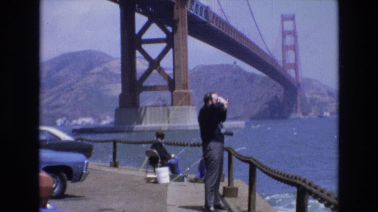 1973 San Francisco waterfront frame showing a man looking through binoculars on a concrete pier, with a seated fisherman, blue car, bay water, hills, and the Golden Gate Bridge in the background.