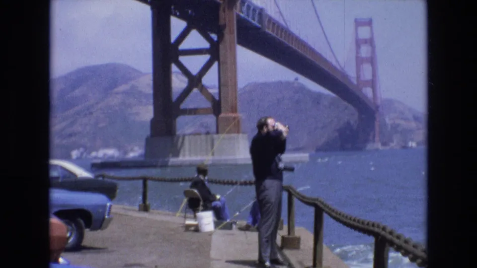 1973 San Francisco waterfront frame showing a man looking through binoculars on a concrete pier, with a seated fisherman, blue car, bay water, hills, and the Golden Gate Bridge in the background.