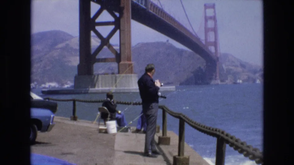 1973 San Francisco home-movie frame showing a man on a concrete pier after using binoculars, a seated fisherman with a bucket, a blue car at the edge of frame, bay water, hills, and the Golden Gate Bridge.