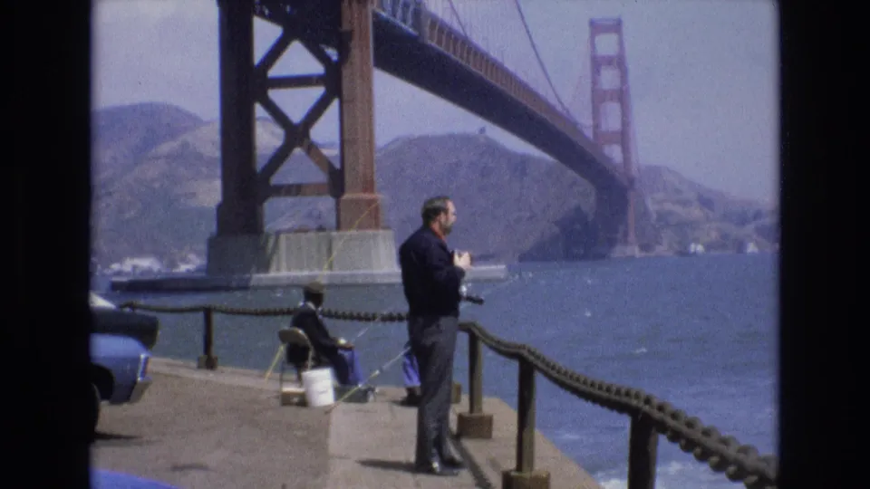 1973 San Francisco waterfront frame with a standing man near the railing, a seated fisherman beside a white bucket, a partial blue car, bay water, distant hills, and the Golden Gate Bridge overhead.