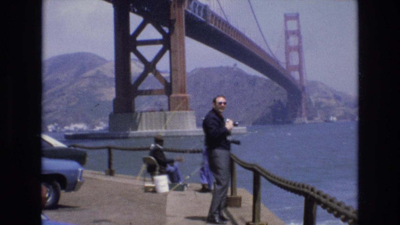 In 1973 San Francisco, a man in sunglasses stands near a rope railing on a waterfront pier, with the Golden Gate Bridge, bay, hills, a seated fisher, and part of a blue car visible.