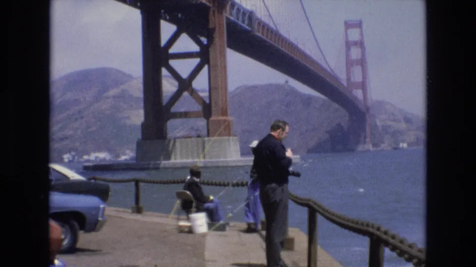 A 1973 waterfront view shows an adult visitor checking a camera beside a rope railing, while another person sits fishing near the bay under the Golden Gate Bridge.