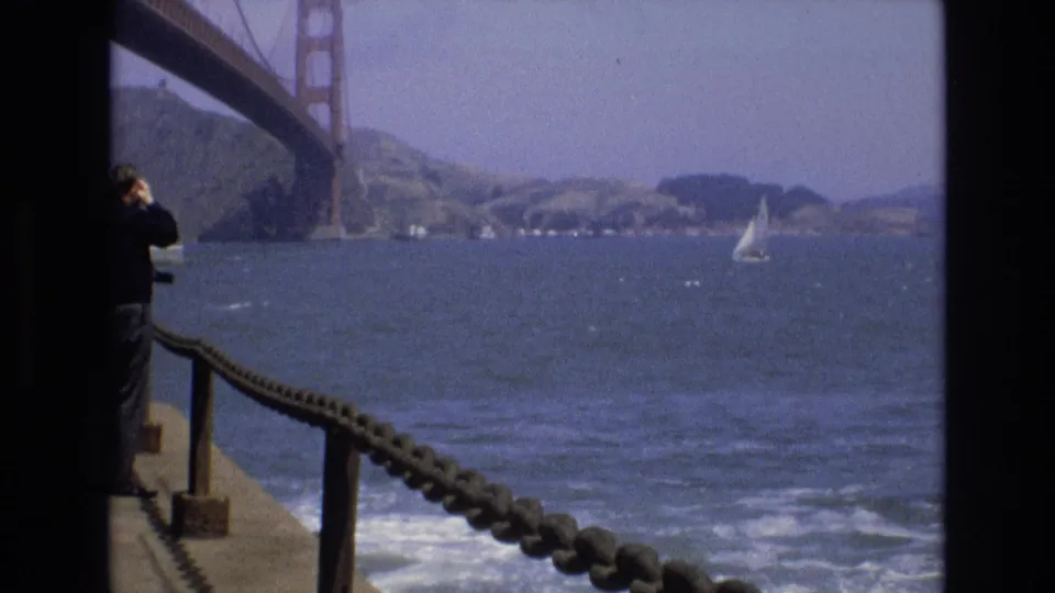 From the San Francisco pier, the frame looks past a partly visible adult visitor and rope railing toward choppy bay water, the Golden Gate Bridge, distant shoreline, and a small white sailboat.
