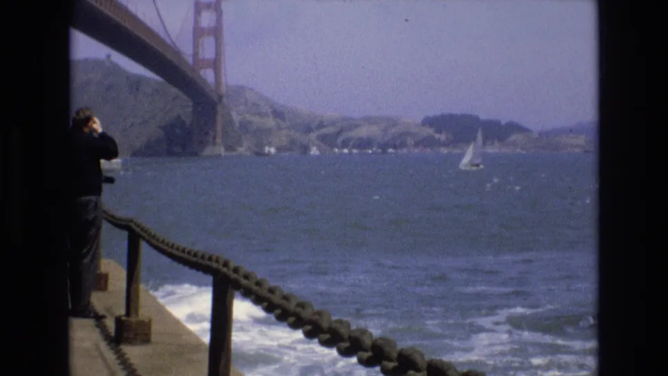 The final 1973 San Francisco frame holds on the bay below the Golden Gate Bridge, with a white sailboat on the water, shoreline in the distance, rope railing in front, and an adult visitor partly cropped at left.