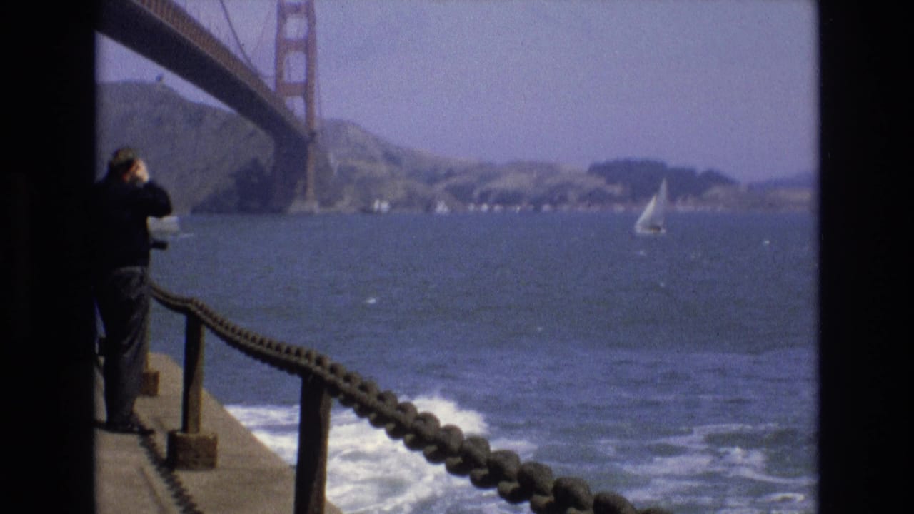 In 1973 San Francisco home-movie footage, a man stands by a rope railing on a pier and looks through binoculars toward the Golden Gate Bridge, bay water, shoreline hills, and a small sailboat.