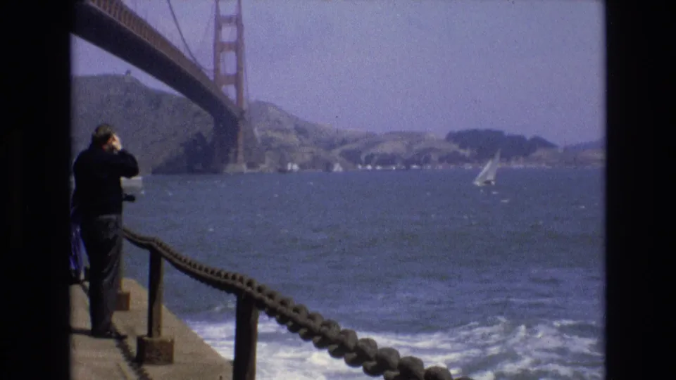 A 1973 San Francisco waterfront frame shows a man at the left edge using binoculars beside a rope railing, with the Golden Gate Bridge overhead and a sailboat crossing the bay.