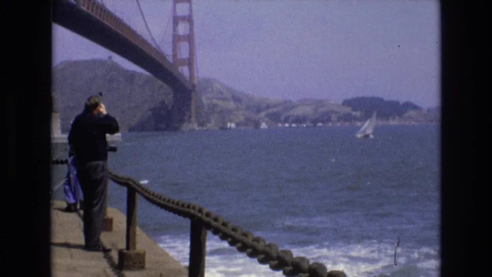 The 1973 8mm frame shows a man in dark clothing at a San Francisco pier railing, looking through binoculars toward the Golden Gate Bridge, choppy bay water, a sailboat, and distant shoreline.