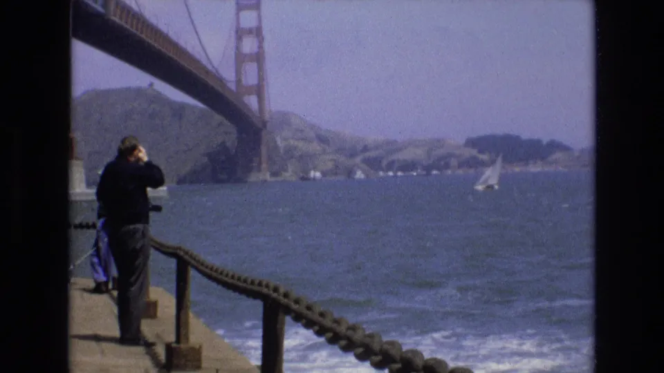 In this supported 1973 San Francisco frame, the Golden Gate Bridge crosses the upper left while a man watches from a pier with binoculars, a small sailboat moves across the bay, and shoreline hills sit in the distance.