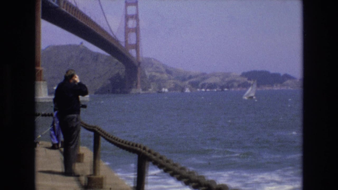 1973 San Francisco home-movie frame showing a man at a bayfront railing using binoculars, with the Golden Gate Bridge, water, a sailboat, and hills in the background.