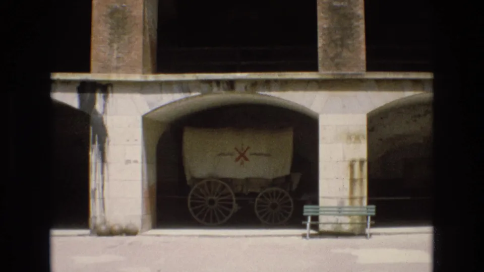 Home-movie frame from San Francisco showing a covered wagon display with a red cross-style emblem under an arched stone structure, with a green bench nearby.
