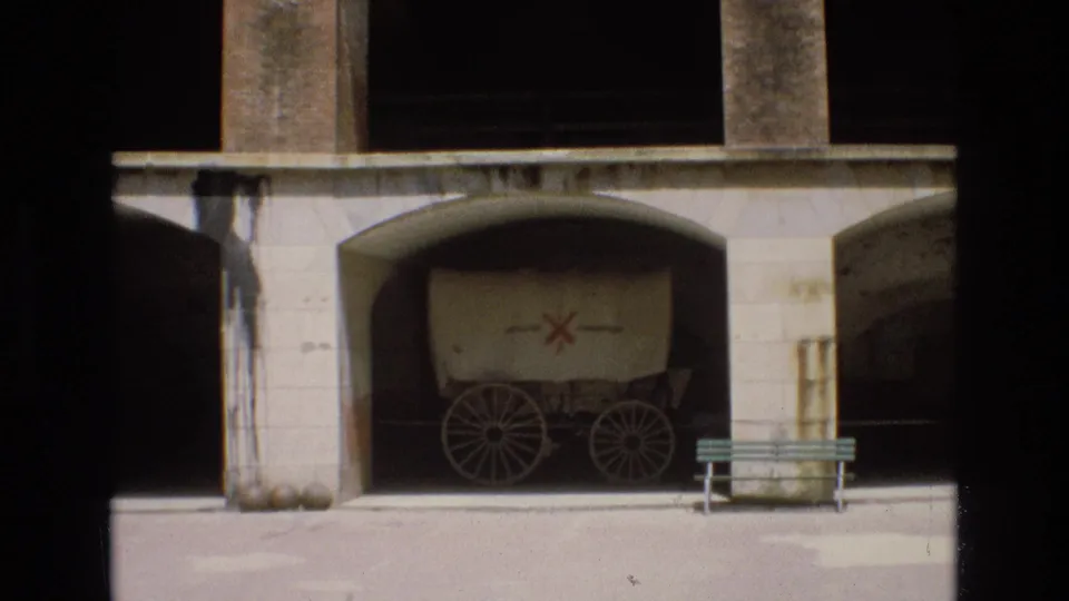 1973 8mm frame showing a covered wagon display beneath a stone arch, with cannonballs near the wall and a green bench on the paved foreground.