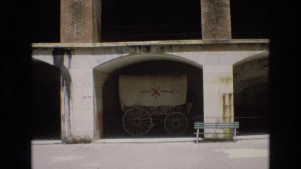 Home-movie frame showing the same covered wagon display under an arched stone opening, with the red cross-style emblem, wagon wheels, bench, and surrounding stonework visible.
