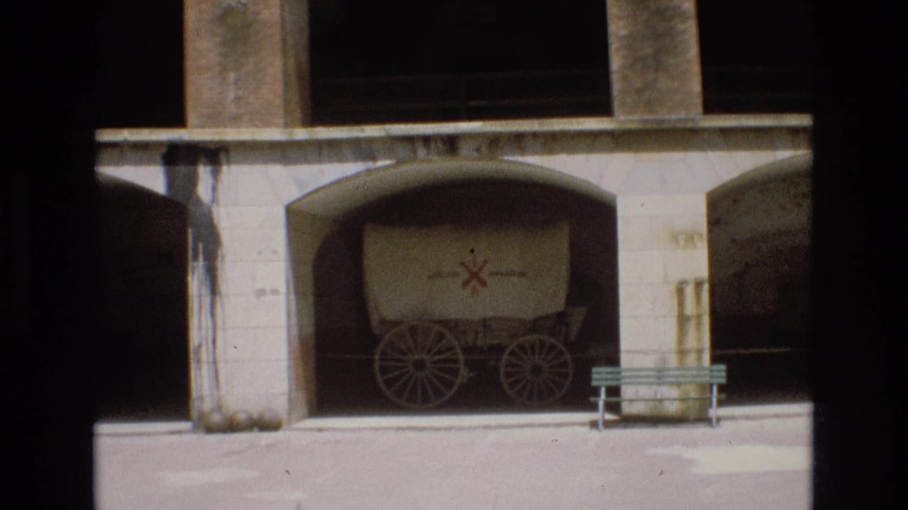 1973 San Francisco 8mm frame showing a covered wagon with a red cross-like marking parked under a concrete archway, with a bench and barrel nearby.