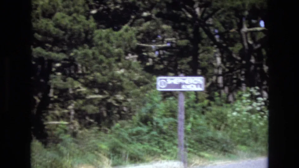 1973 San Francisco 8mm frame showing a roadside sign that appears to read Shepherd's Knoll, surrounded by trees, shrubs, and a pale road edge.
