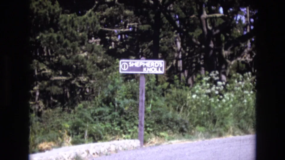 1973 San Francisco 8mm frame with the Shepherd's Knoll sign centered against wooded foliage and a light roadside surface in the foreground.