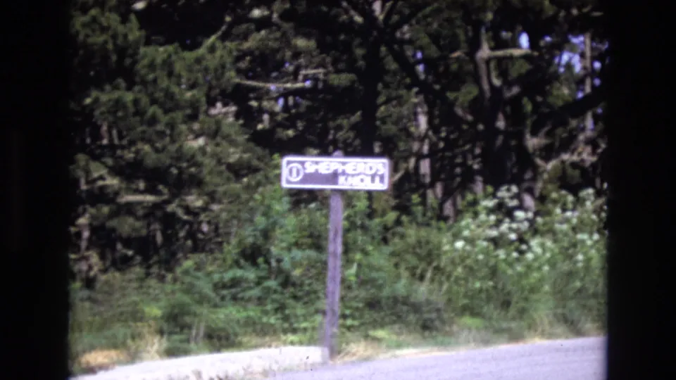 1973 San Francisco 8mm frame showing the Shepherd's Knoll sign slightly left of center, with a curb, roadside vegetation, and dark trees behind it.