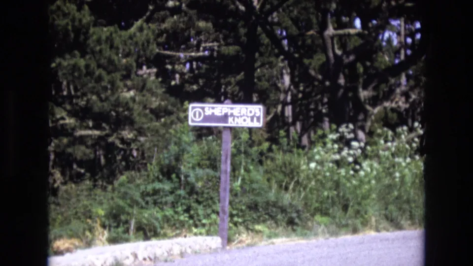 1973 San Francisco-area 8mm frame showing a small wooden Shepherd's Knoll sign on a post near a road shoulder, backed by shrubs and tall trees.
