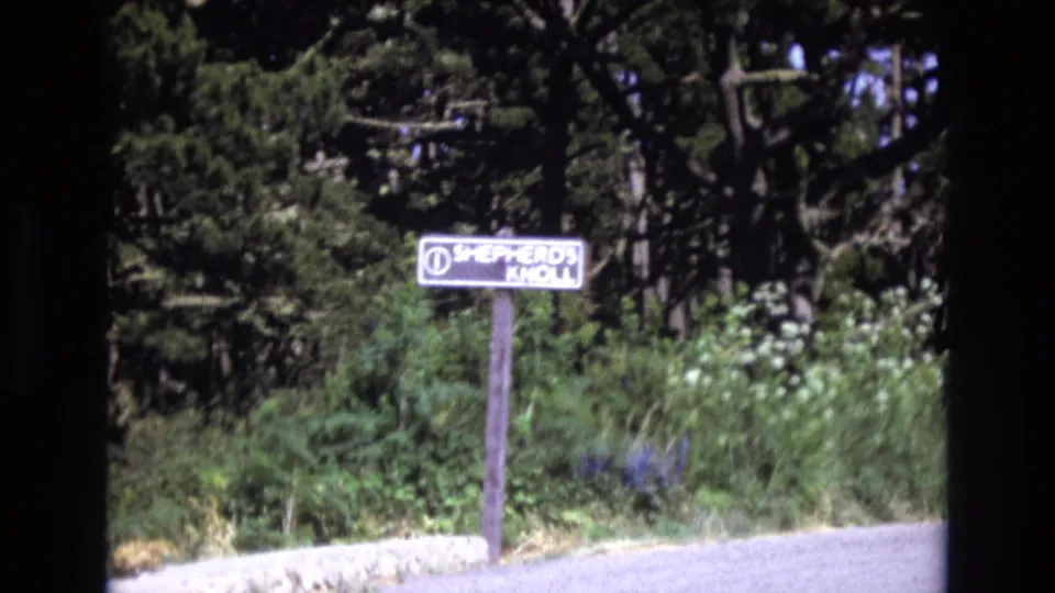1973 San Francisco-area home-movie frame with the Shepherd's Knoll sign centered above overgrown foliage beside a quiet road.