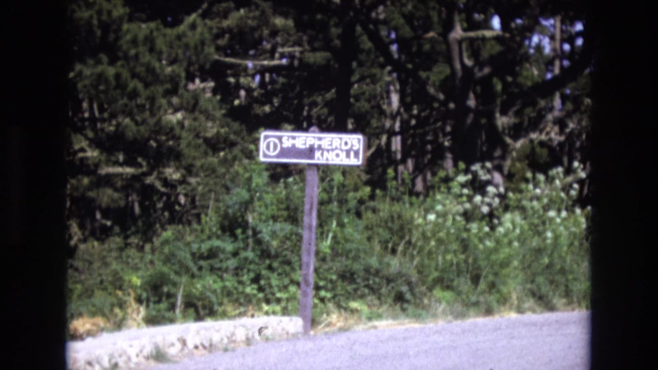 Weathered Shepherd's Knoll sign beside a path with shrubs and conifer trees behind it.