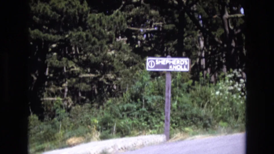 1973 San Francisco 8mm frame showing the Shepherd's Knoll sign slightly lower in the frame, with a stone curb, pale path, thick shrubs, and conifer trees behind it.