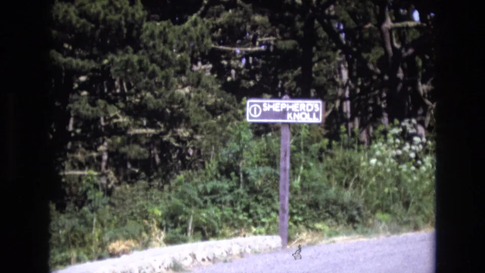 1973 San Francisco 8mm frame holding on the Shepherd's Knoll sign next to a pale path, with dense vegetation, conifer branches, and visible film-frame borders.