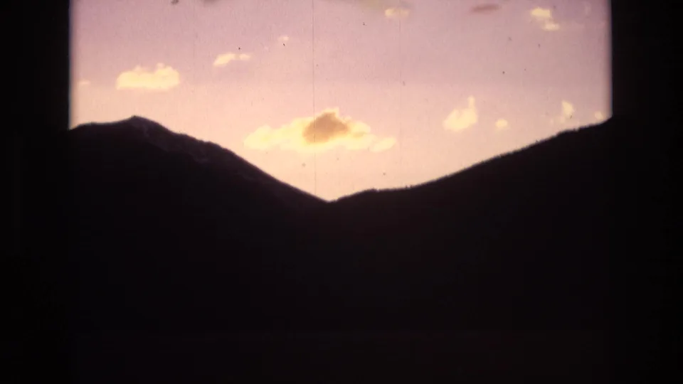 A shadowed Colorado Springs mountain range fills the lower frame while a glowing orange-yellow horizon and scattered clouds sit above it in grainy 8mm color.