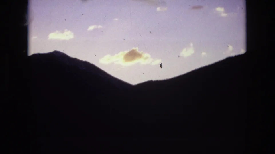 The final 1974 8mm frame shows a dark mountain silhouette near Colorado Springs under a cooler blue-lavender sky, with pale clouds and visible film specks.