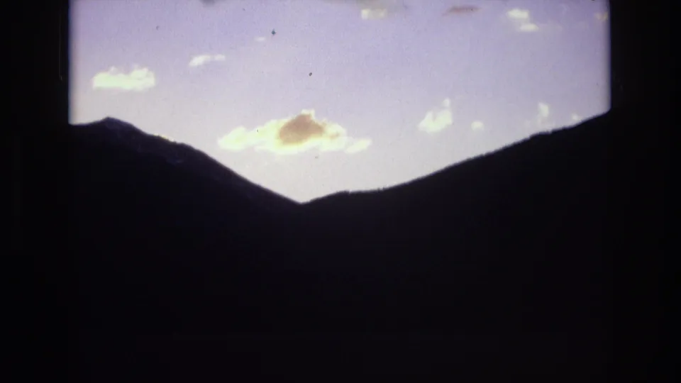 1974 home-movie frame from Colorado Springs showing a mountain valley in deep shadow while dawn light touches clouds above the ridge.