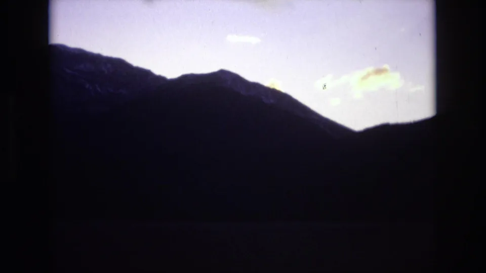 1974 Colorado Springs home-movie frame showing a dark mountain silhouette under a pale lavender dusk sky with faint glowing clouds.