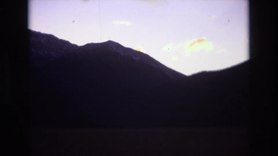 1974 Colorado Springs home-movie frame showing dark mountain slopes, a central ridge dip, and small pale clouds in a faded dusk sky.