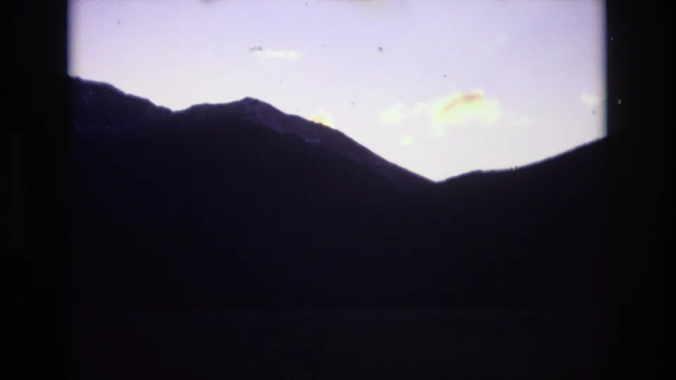 1974 Colorado Springs 8mm frame holding on a dark mountain silhouette, pale evening sky, small clouds, and heavy shadow across the lower frame.