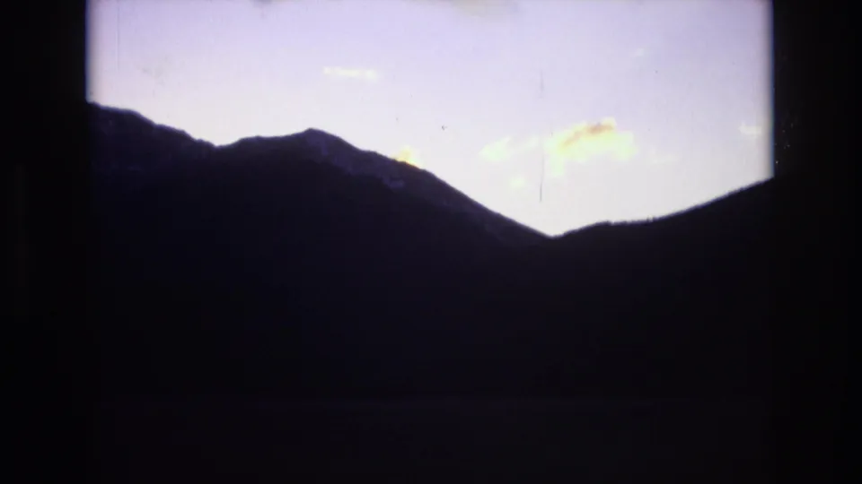 A dark mountain slope and valley form a heavy silhouette under a pale evening sky in the 8mm Colorado travel footage.