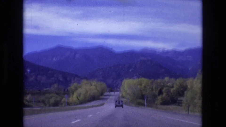 A two-lane road runs through trees toward distant blue mountain ranges, with a small car visible ahead in the 1974 Colorado Springs road-trip footage.
