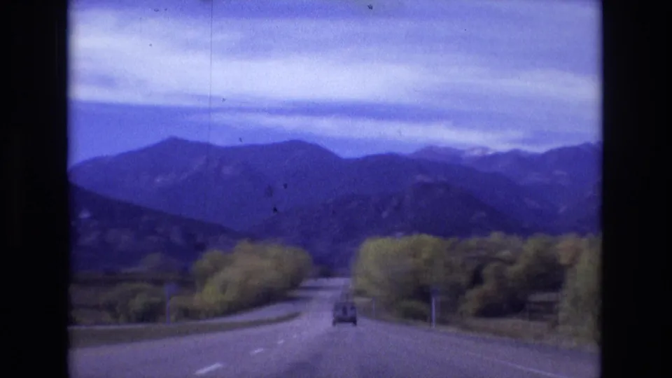 1974 Colorado Springs area home-movie frame showing a vehicle ahead on a paved mountain road bordered by trees, with blue-toned mountains and clouds in the distance.