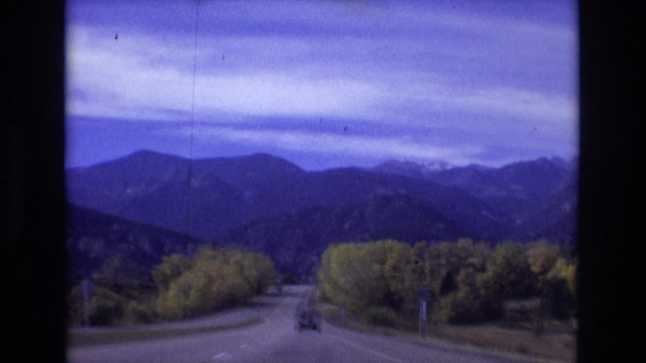 A vehicle follows a curving road with trees and mountains under a cloudy sky.