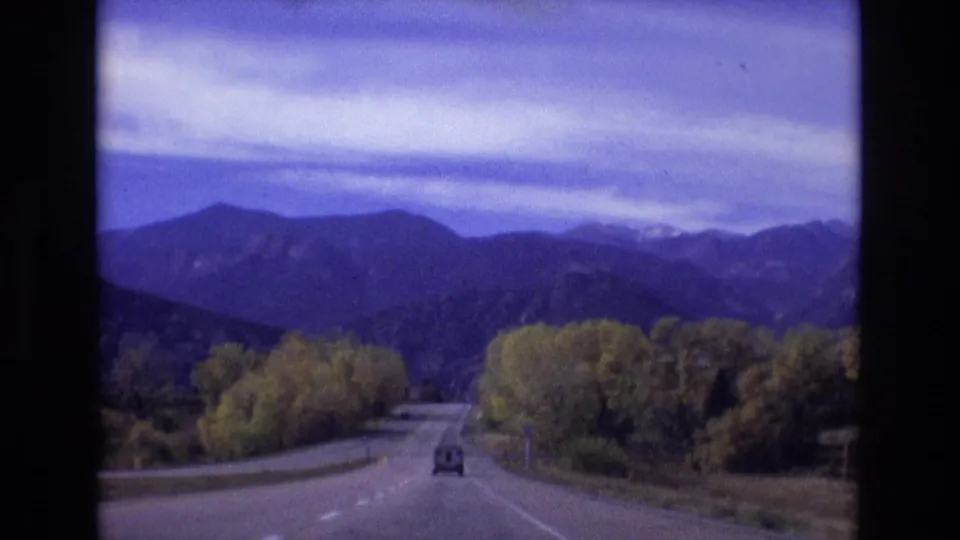 1974 Colorado Springs area frame from a moving vehicle, showing a small vehicle ahead on a winding road lined with trees and backed by dark blue mountains.